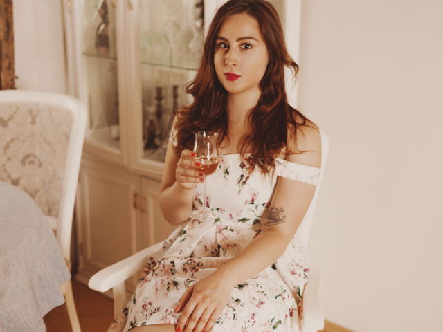 ViaLattea with wavy brown hair, red lipstick, floral dress, holding a glass of wine, tattoo on right arm, sitting, white chair, wooden floor, soft light, relaxed expression.