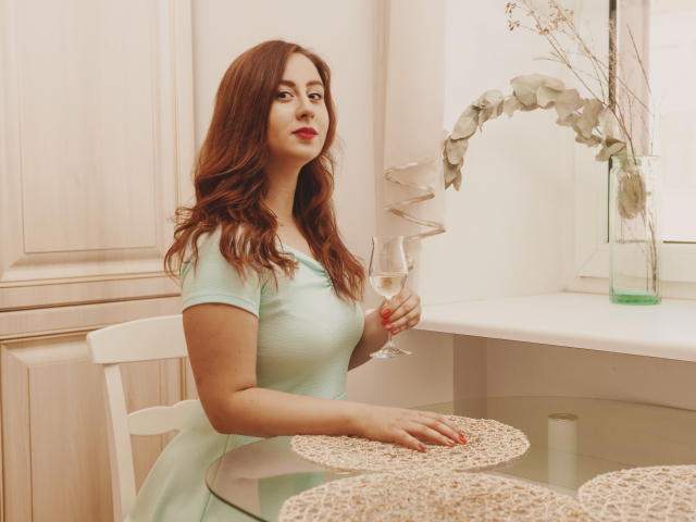 ViaLattea with red hair, light green dress, holding a champagne flute, sitting at a table with woven placemats, white chair, and ornamental plant.
