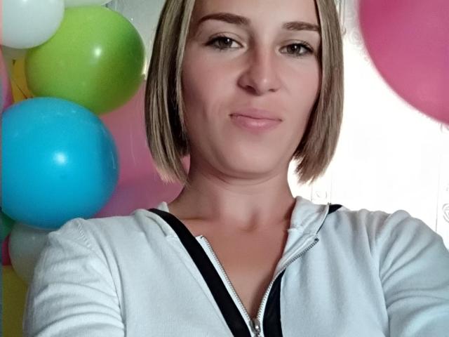 SueShine ha i capelli biondi corti, la pelle chiara e un seno piccolo, indossa una camicia bianca con zip, ha un leggero sorriso ed è circondata da palloncini colorati..