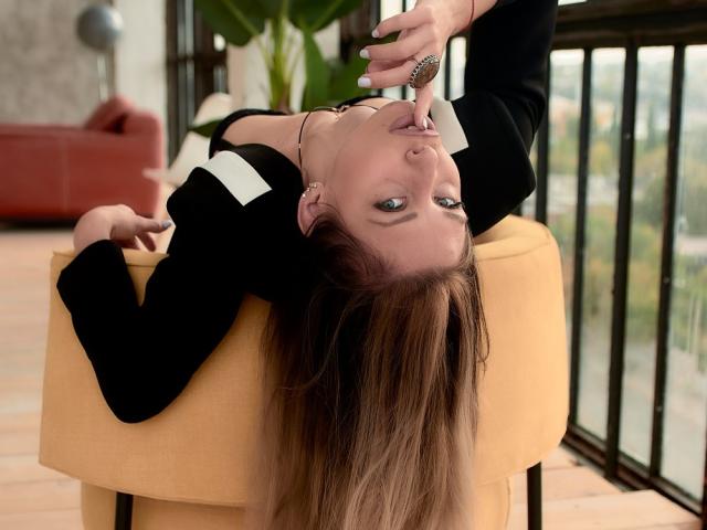 MisissFire, upside-down on a yellow chair, black blazer, white collar, long brown hair, finger in mouth, looking at camera, ring on finger, indoor, natural light, casual.