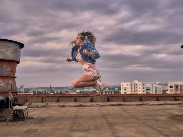 MisissFire is mid-air, jumping on a dirty rooftop, wearing a denim jacket and pink shorts, with a cloudy sky and rusty barrel in the background.
