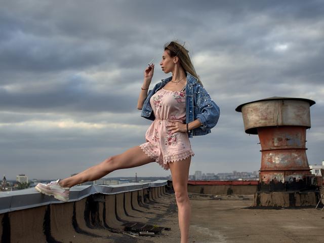 MisissFire with a slim, sexy body, pink floral romper, and blue denim jacket, standing on a rooftop, leg up, smoking, white sneakers, cloudy sky, rusted water tower.