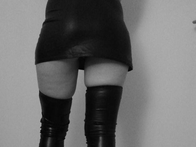 HotJolieX indossa una minigonna in pelle nera, mostrando il suo sedere scoperto, con stivali neri alti fino alla coscia, in una fotografia in bianco e nero. Le sue cosce sono leggermente divaricate..