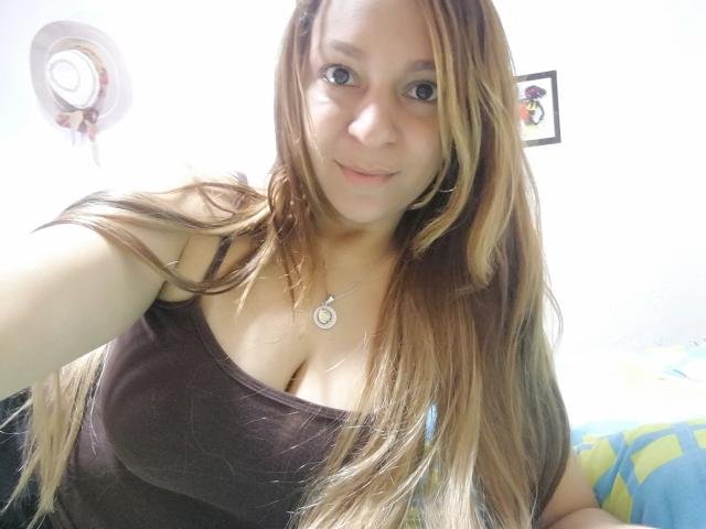 Angelonix with long brown hair, wearing a brown tank top, showing her big tits, small necklace, and smiling at the camera. She has light skin and blue eyes.