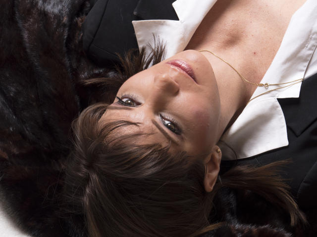 MistressLaurie's face with dark brown hair, green eyes, and subtle makeup, lying on a black fur, wearing a white shirt and black suit, with a gold necklace.
