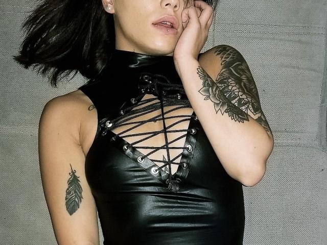 KassiKassou met een zwarte leren top, vetergesloten borst, tatoeages op de armen en onderarmen, hand in de buurt van de mond, kort zwart haar, lichte huid, kleine borsten, zichtbare tatoeages, grijze achtergrond..