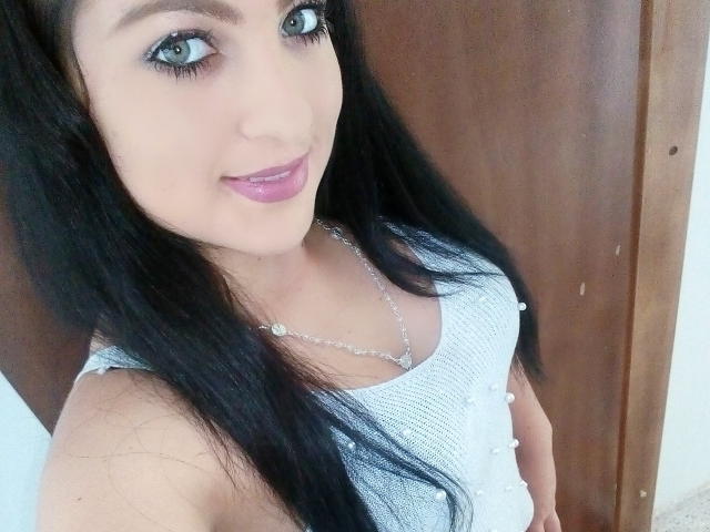 Evelyn Fox, con lunghi capelli neri, occhi verdi e rossetto viola, indossa una canotta bianca e una collana d'argento, sorride alla fotocamera, mostrando il seno..