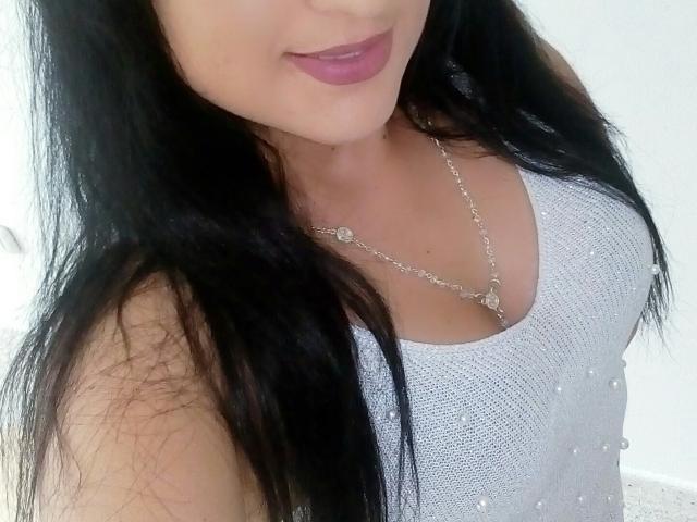 Evelyn Fox, con lunghi capelli neri, indossa una canottiera grigia scintillante che mette in mostra il suo seno piccolo, una collana d'argento e rossetto viola, in un selfie..