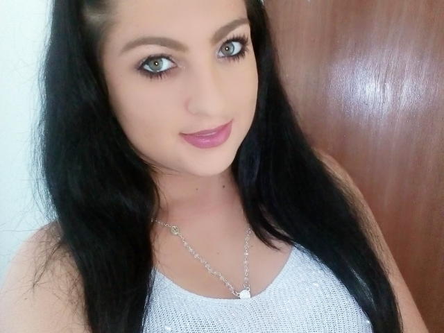 Evelyn Fox, avec de longs cheveux noirs, des yeux verts, du rouge à lèvres rose, portant un collier en argent et un haut en maille blanche qui laisse entrevoir légèrement ses seins. Elle sourit à l'appareil photo..
