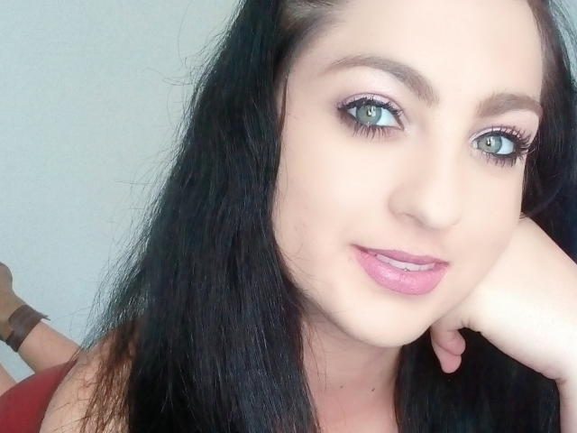 Evelyn Fox hat eine blasse Haut, schwarzes Haar, blaue Augen, pinken Lippenstift und legt ihr Kinn in ihre Hand. Sie trägt ein braunes Armband. Brust nicht sichtbar..