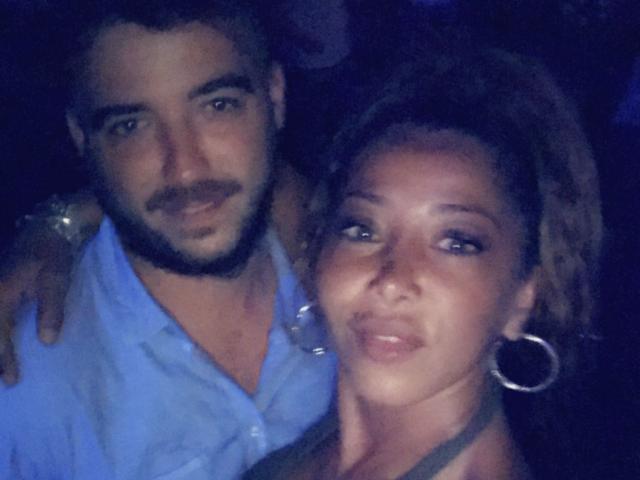 Saraby, com cabelos cacheados escuros e brincos de argola, olhando para a câmera, está ao lado de Saraby, com barba e camisa azul, ambos sorrindo. Fundo escuro..