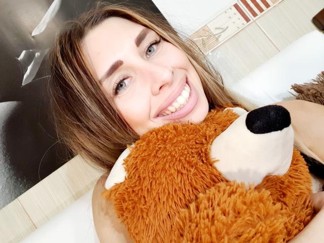 MisissFire with long brown hair, smiling, holding a fluffy orange teddy bear, showing white teeth, in a bed, with a black and white wall poster partially visible.