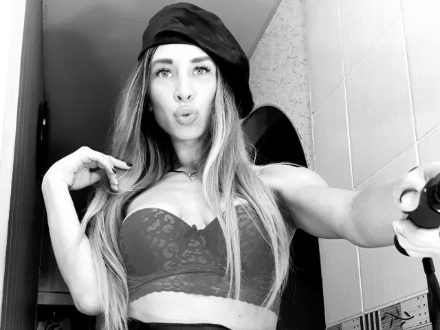 MisissFire with long hair, wearing a baseball cap and lace bra, pouting lips, holding phone, showing her tits, with a ring on her finger, in a black and white selfie.