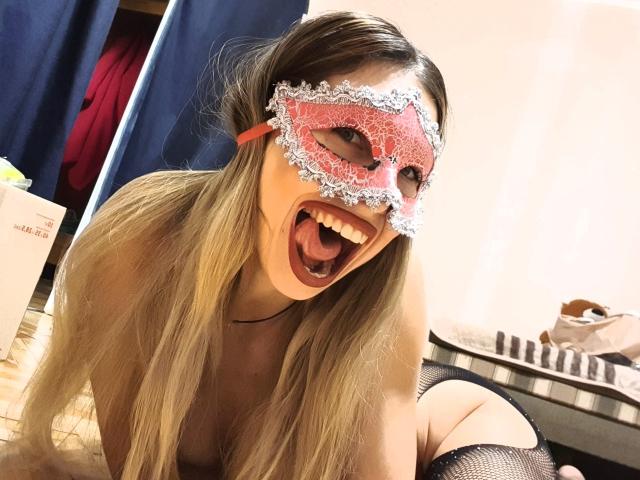 MisissFire with long blonde hair, wearing a red lace masquerade mask with silver frills, open-mouthed, smiling, kneeling, topless, black fishnet stockings, in a bedroom.