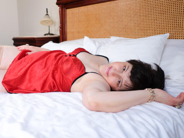 ChristaRose lies on a bed, wearing a red satin nightie that shows her small tits, with her arms above her head, looking at the camera with pink lipstick.