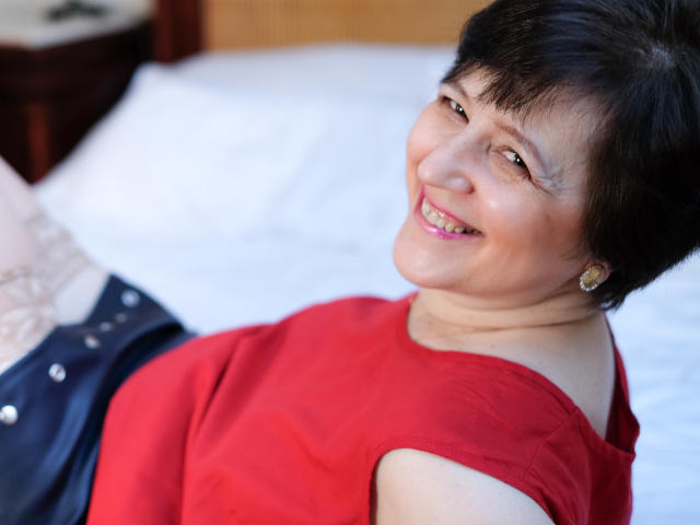 ChristaRose, short black hair, smiling, red off-shoulder top, small round earrings, lying on white bed, navy skirt with button details, fair skin, slightly chubby, pink lipstick.