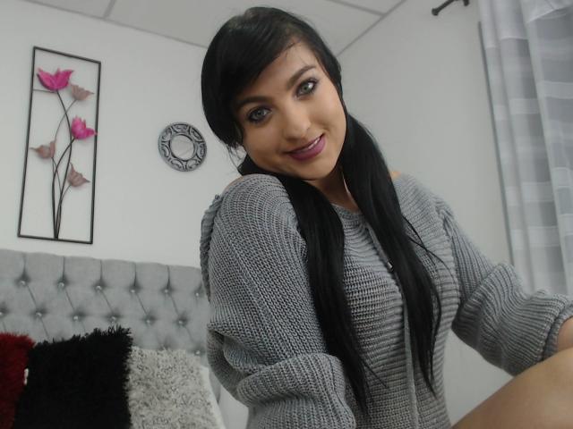 EvelynFox, avec ses longues nattes noires, son pull gris en tricot et son rouge à lèvres violet, sourit à l'appareil photo, montrant légèrement sa poitrine. Chambre avec une tête de lit grise capitonnée et des décorations murales florales..