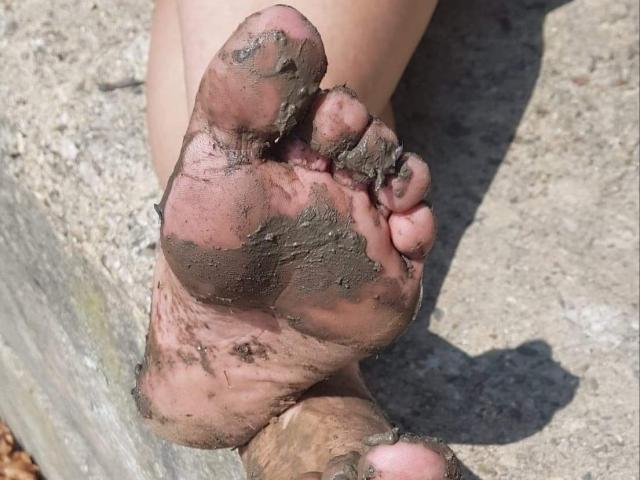 LaDominatrice's dirty, mud-covered sole is in the foreground, with her toes slightly curled and her heel resting on a wooden surface. Her foot is dirty and pinkish.