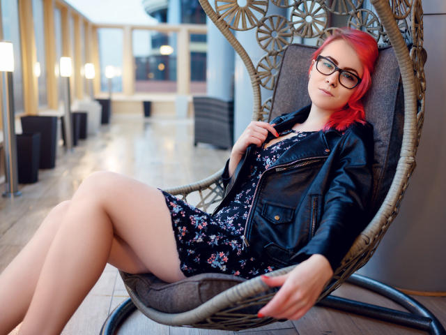 Lilian Kroft, con cabello rojo, gafas, chaqueta de cuero negro, vestido floral y piernas desnudas, sentada en una silla de mimbre, con las piernas separadas, las manos sobre la chaqueta, mirando a la cámara..