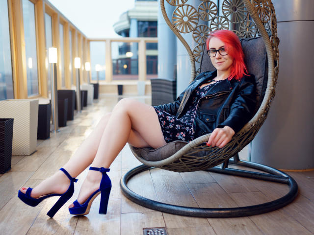 LilianKroft with red hair, glasses, black leather jacket, floral dress, and blue high heels, sitting in a wicker chair, legs crossed, smiling, wooden floor, modern setting.