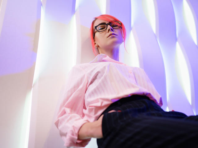 LilianKroft with red hair, black glasses, pink striped shirt, hands in black pants, looking down, purple and white lighting, low angle shot.