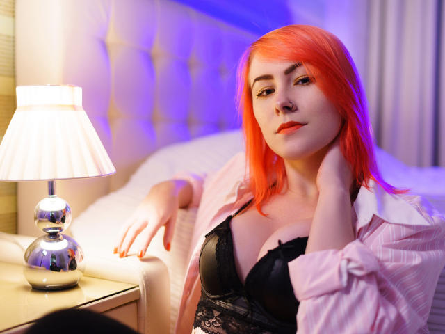 LilianKroft with bright red hair, wearing a black lace bra and pink shirt, sitting by a bed with a lamp, her hands near her neck, looking at the camera.
