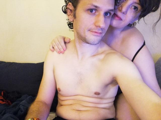 LolaAndMike, avec ses cheveux bouclés, ses petites seins et son bandeau noir, enlace la poitrine nue, légèrement ronde et pâle de Mike. Les yeux bleus de Mike sont détendus, et le visage de Lola est près de son épaule..