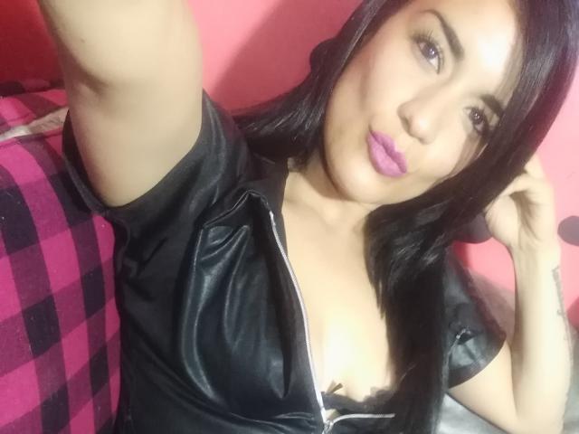 ValeryGirlLatin met zwart haar, paarse lippenstift, een zwarte leren top en zichtbare decolleté, met arm omhoog, kijkend naar de camera, op een roze en zwarte geruite bank..