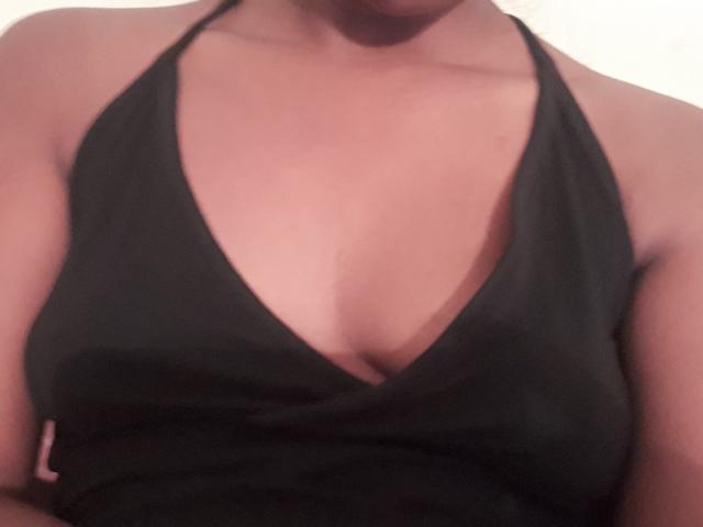 La blusa negra de halter de SexyPoosy tiene un escote en V pronunciado, que muestra sus senos de tamaño medio, bronceados y desnudos. La tela de su blusa está ligeramente arrugada. La imagen es una fotografía con enfoque suave..