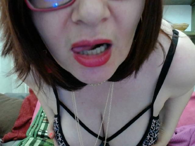 BigTitsXHot with red lipstick, glasses, and a leopard print bra, leaning forward, showing cleavage, and tongue slightly out, wearing a gold necklace.