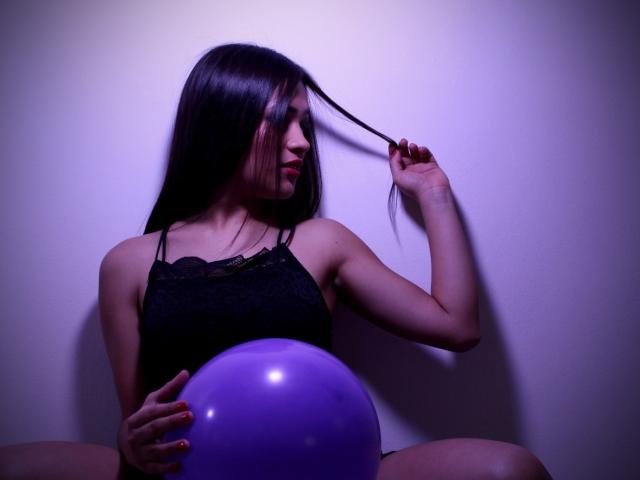 CelesteEvanss with long black hair, holding a purple balloon, wearing a black lace top, her hand in her hair, sitting with legs spread, in dim purple lighting.