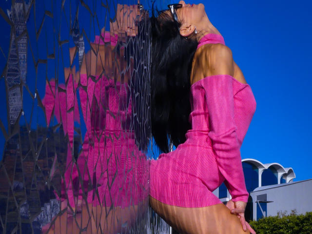 CassandraMichelli with long black hair, wearing a pink off-shoulder knit dress, leaning against a reflective glass wall, showing her ass and reflecting her face and body.