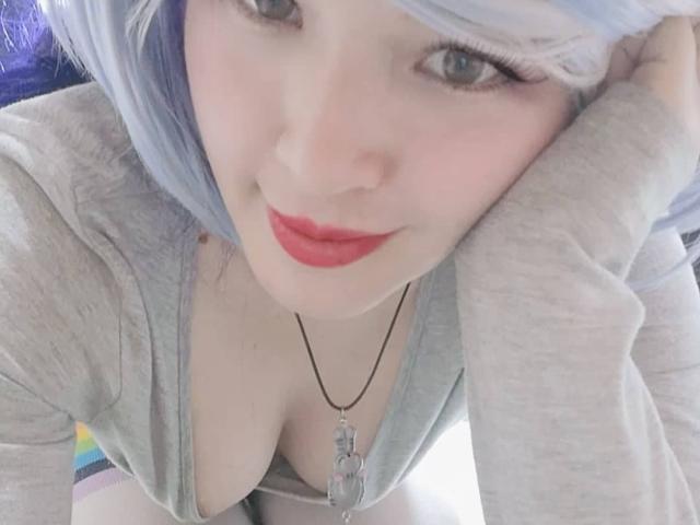 HikaruChan met lichtblond haar, rode lippenstift en een grijze top, leunt voorover, waarbij haar decolleté zichtbaar is, draagt een ketting met een transparante hanger en glimlacht naar de camera..