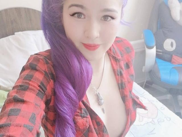 HikaruChan avec des cheveux violets, une chemise à carreaux rouges, décolletée, révélant sa poitrine, du rouge à lèvres rouge et un eye-liner noir, souriant à la caméra, un collier avec un pendentif, assise sur un lit..