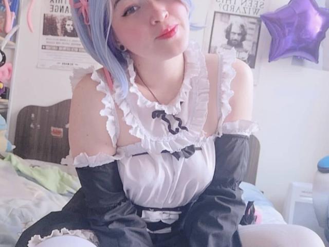 HikaruChan mit blauen Haaren, die ein schwarz-weißes, gerauschtes Dienstmädchenkleid trägt, lächelt mit roten Lippen, sitzt auf einem Bett und zeigt weiße, hohe Strümpfe. Sie ist süß und sexy..
