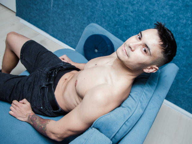 MattMalone, senza camicia, con un corpo asiatico tonico e muscoloso, capelli neri corti, tatuaggio sull'avambraccio destro, pantaloncini neri, sdraiato su un cuscino blu, che guarda la fotocamera, con un'espressione rilassata..