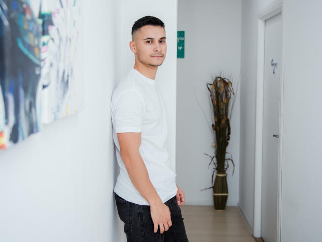 MattMalone, debout dans un couloir aux murs blancs, portant un t-shirt blanc et un pantalon noir, avec des cheveux noirs courts, regardant par-dessus son épaule, dans une pose détendue et confiante..