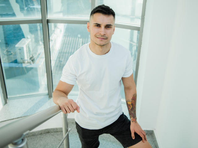 MattMalone with a white t-shirt, black shorts, and a colorful tattoo on his right arm, leaning on a railing, smiling, short dark hair, standing in a sunlit room.