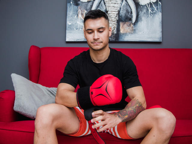 MattMalone, gespierd, zwart T-shirt, rode bokshandschoenen, oranje korte broek, getatoeëerde arm, kort donker haar, rode bank, zittend, ogen gesloten, rechterhand op de kruis, linkerhand op de handschoenen..