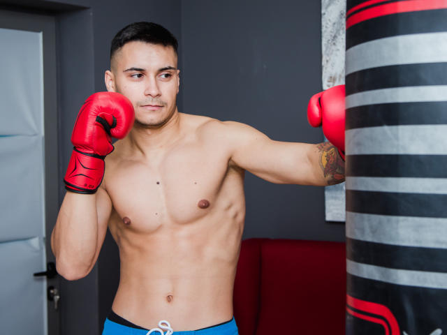 MattMalone, sin camisa, con guantes de boxeo rojos, un tatuaje en el brazo derecho, pantalones cortos azules, golpeando un saco de boxeo a rayas negras y rojas, tiene un cuerpo musculoso y tonificado y una expresión seria..
