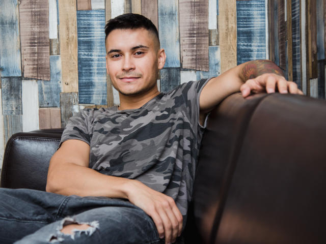 MattMalone mit kurzen, dunklen Haaren, einem Tarn-T-Shirt und zerrissenen Jeans, lehnt an einem braunen Ledersofa, hat ein Tattoo am rechten Arm, ein leichtes Lächeln, wirkt lässig und entspannt, der Arm ruht auf dem Sofa..
