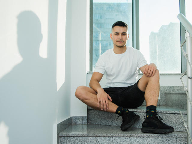 MattMalone, sitting on gray stone steps, wearing a white tee, black shorts, black socks, and black shoes, with a shadow on the white wall to the left.