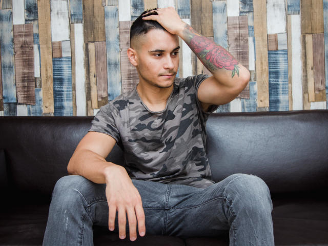 MattMalone, con i capelli neri e corti, indossa una camicia mimetica e jeans blu, è seduto su un divano in pelle nera, con una mano tra i capelli, mostrando un tatuaggio colorato sul braccio..