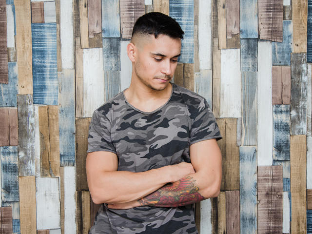 MattMalone, muskulös, tätowierter Arm, trägt ein Tarnhemd, kurze schwarze Haare, Arme verschränkt, blickt nach unten, Hintergrund mit Holzmaserung, ernster Gesichtsausdruck..