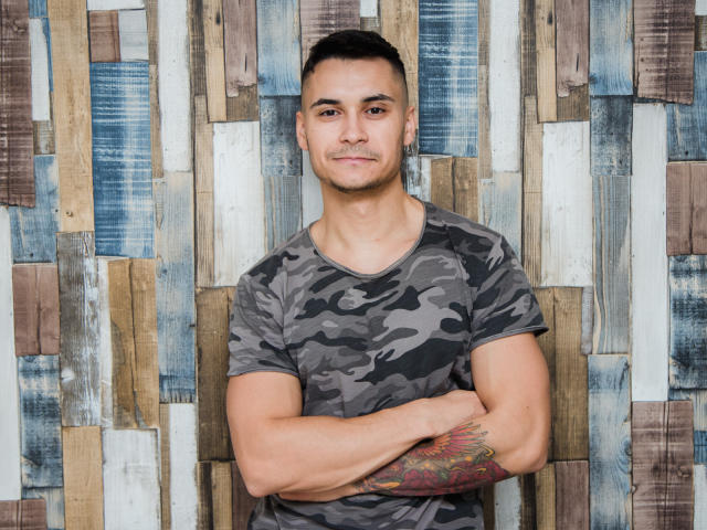 MattMalone, muskulös, trägt ein graues Tarnhemd, Arme verschränkt, Tattoo am rechten Unterarm, kurze schwarze Haare, kleine Ohrringe, steht, lächelt leicht, vor einer Wand aus Holzbrettern..
