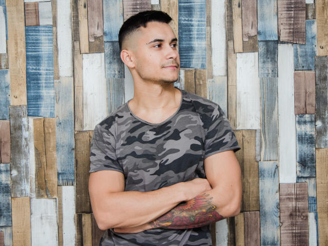 MattMalone, muscular with tattooed arm, wearing camo shirt, arms crossed, short dark hair, looking to the side, slight smirk, standing against wooden patterned wall.