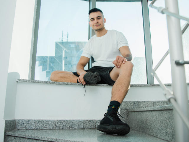 MattMalone sitting on gray stairs, wearing a white tee, black shorts, black socks, and black sneakers, holding a sneaker, looking at the camera, short black hair, light skin.