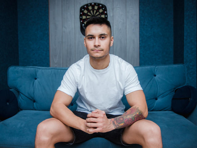 MattMalone, muscular, white tee, black shorts, tattooed right arm, short dark hair, sitting on blue couch, hands on knees, serious expression, dartboard behind him.