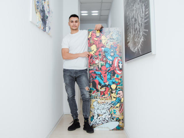 MattMalone se tient debout, tenant un tableau coloré de style bande dessinée avec des super-héros, vêtu d'un t-shirt blanc et d'un jean déchiré, dans un couloir aux murs blancs. Il a une belle silhouette et des cheveux noirs courts..