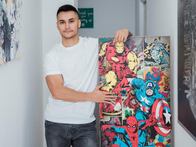 MattMalone with a muscular build, short dark hair, white t-shirt, and blue jeans, holding a colorful comic book featuring superheroes like Spider-Man and Captain America.